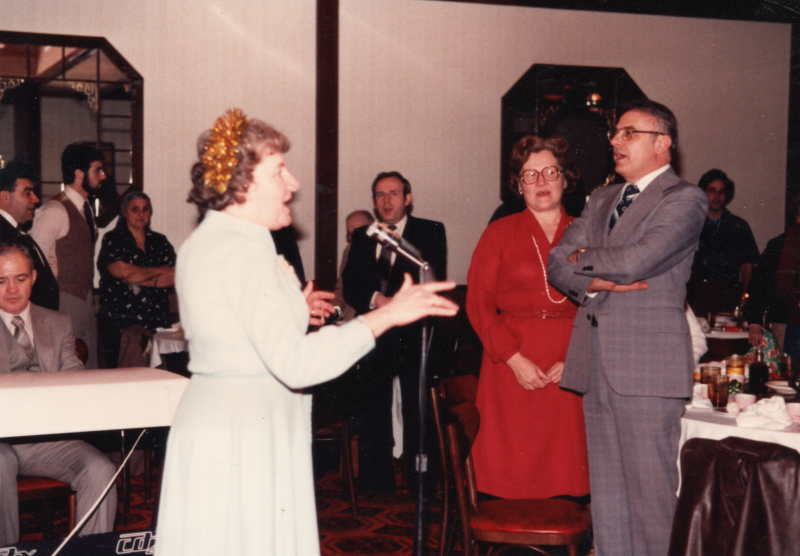 A woman in a blue dress sings into a microphone facing other congrgants singing in a restaurant with a piano accompanist