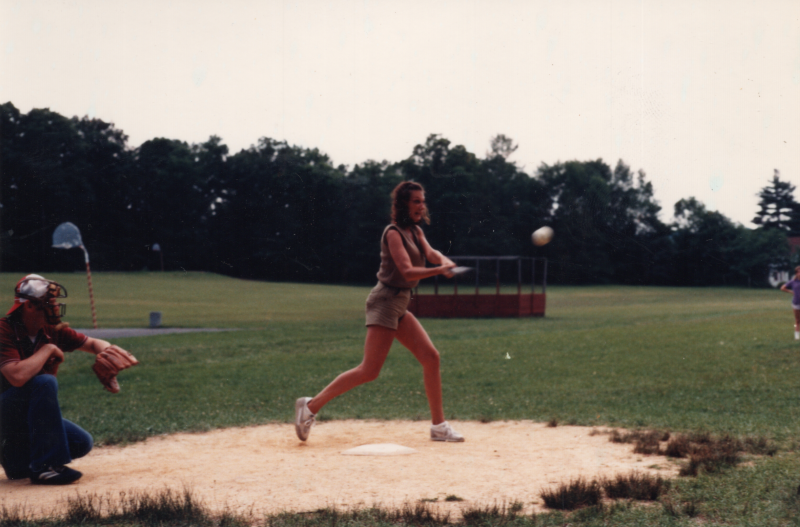 A woman swings a bat to hit a ball on a grassy baseball field