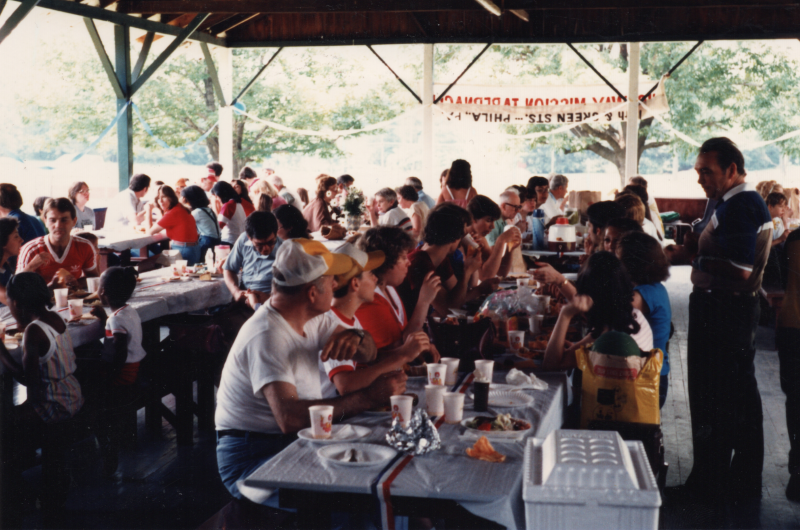 Congregants dine together in an outdoor pavillion with blue and whire decorations and a flipped over sign reading, "Highway Mission Tabernacle, 19th and Green Sts.  . . . Phila., PA"