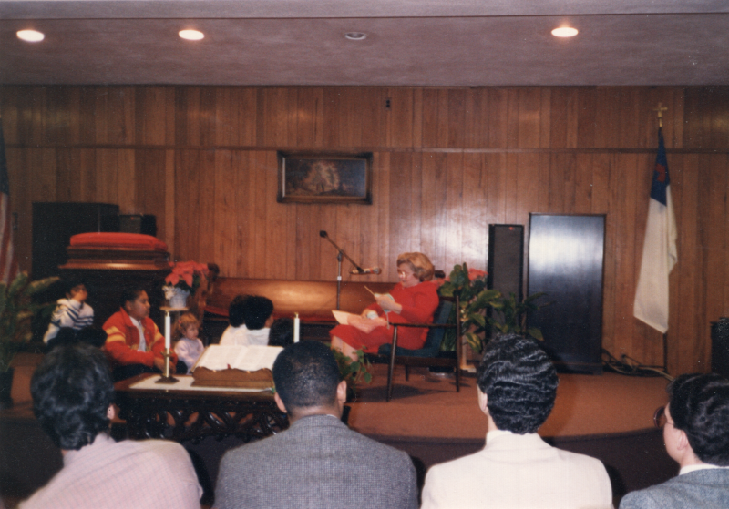 Mrs. Bogdan knits in front of the congregation with a microphone, facing a group of children seated on stage