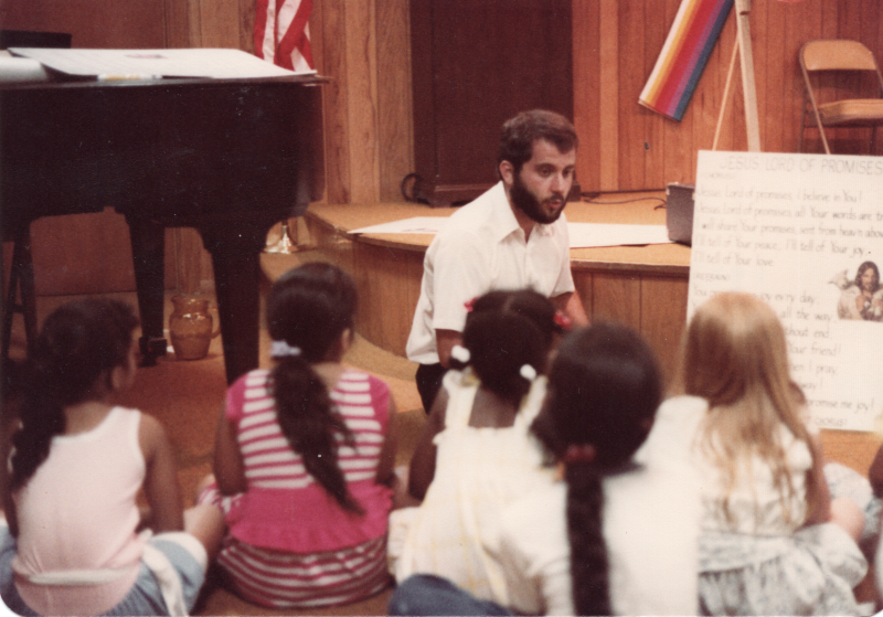 A man speaks to a group of children using a poster titled, "Jesus, Lord of Promises"