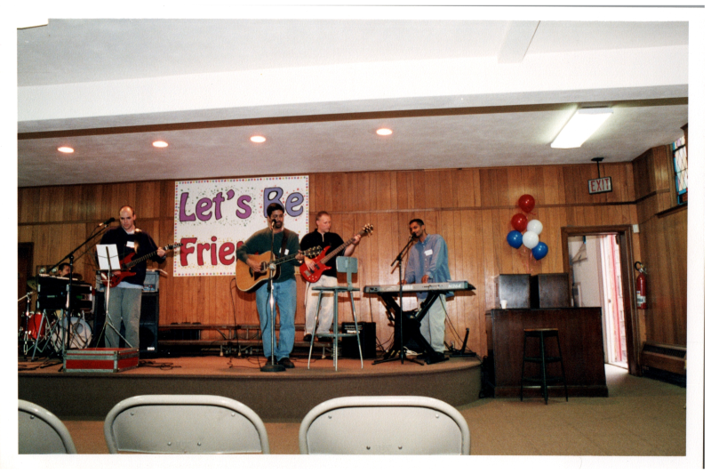A group performs in the back of the church in front of a banner reading "Lets Be Friends!"