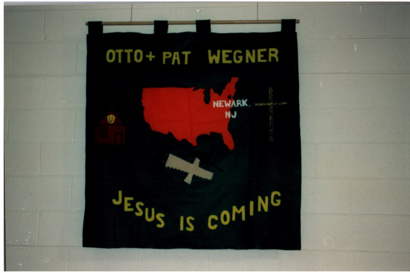 Picture of the Wegners' missions convention banner reading, "Otto + Pat Wegner, Jesus is Coming" with imagery of a church, Newark, NJ, a cross, and construction tools. Caption on the back reads, "1998 Missions Convention, A/G Christian Center, St. Charles, IL, Pastor Gary Blanchard, We are praying for You!"