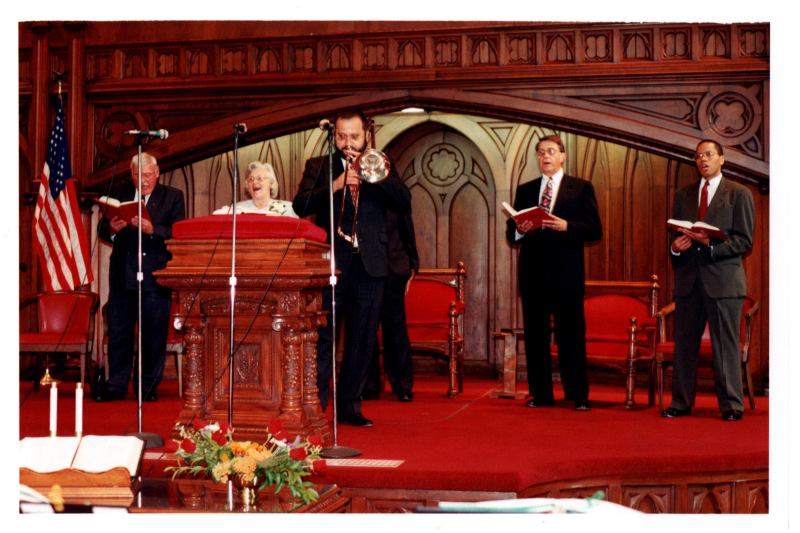 A man plays a trumpet with Otis A. Fortenberry, Otto Wegner, Mrs. Bogdan, and another man singing from hymnals