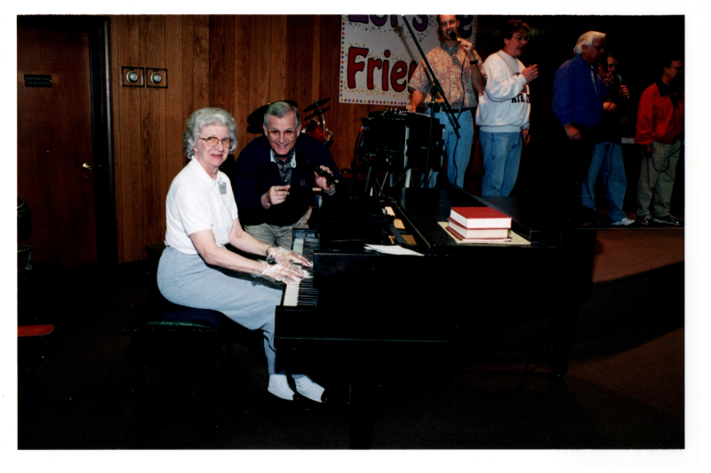Mrs. Betty poses with a man for a photo while playing the piano for a group of singers