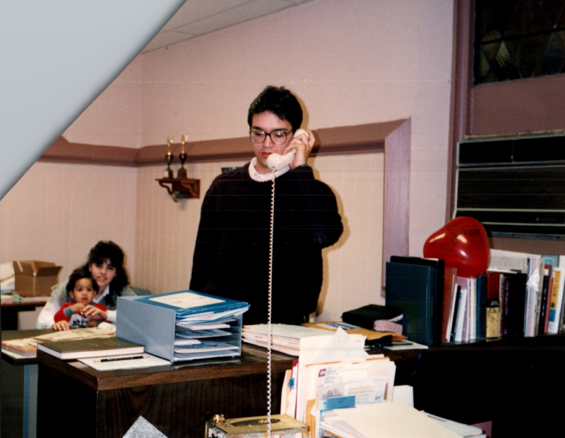 Mark DaSilva answers the phone while Elda holds a baby in the background