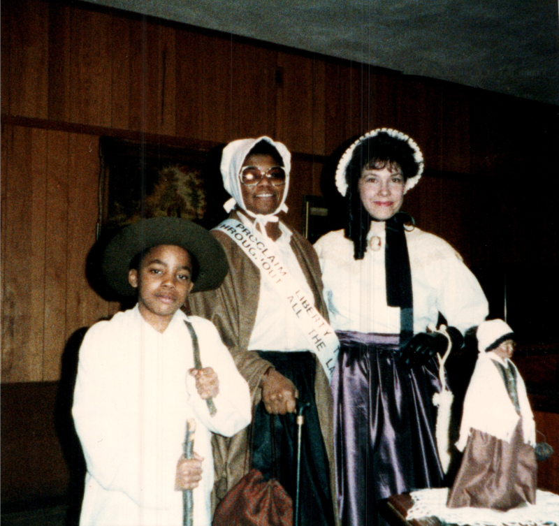 Three people in costume-- a young boy in a hat, an older woman with a bonnet and a sash reading, "Proclaim Liberty through all the land," and a woman in a bonnet-- smile together for a photo