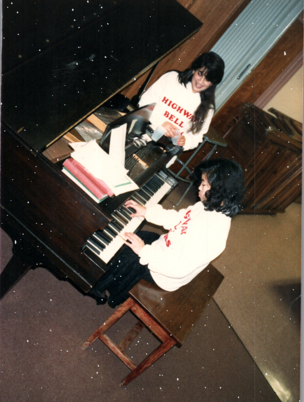 Two women in bell choir sweatshirts play and talk at a piano