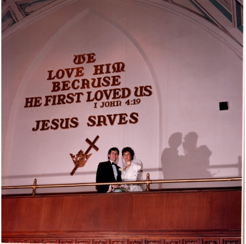 A bride and groom stand together under the sign in the sanctuary reading, "We Love Him Because He First Loved Us, I John 4:19" with the Cross and Crown.