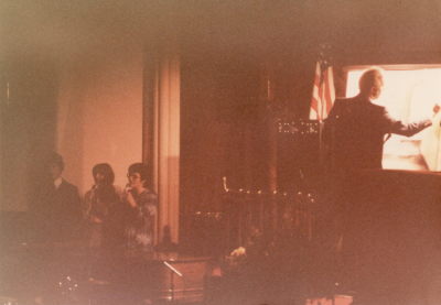 A man draws on an illuminated screen while three peeople sing